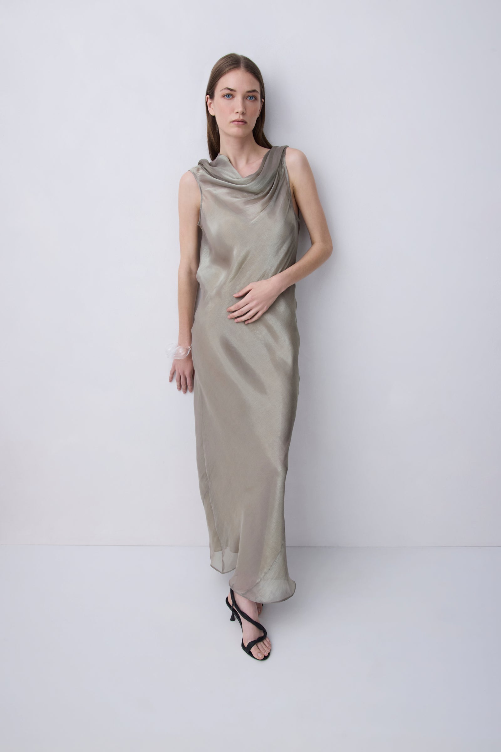 A woman modeling a sleeveless, cowl-neck maxi dress in a shimmering champagne-gold satin, standing against a plain light gray studio background.