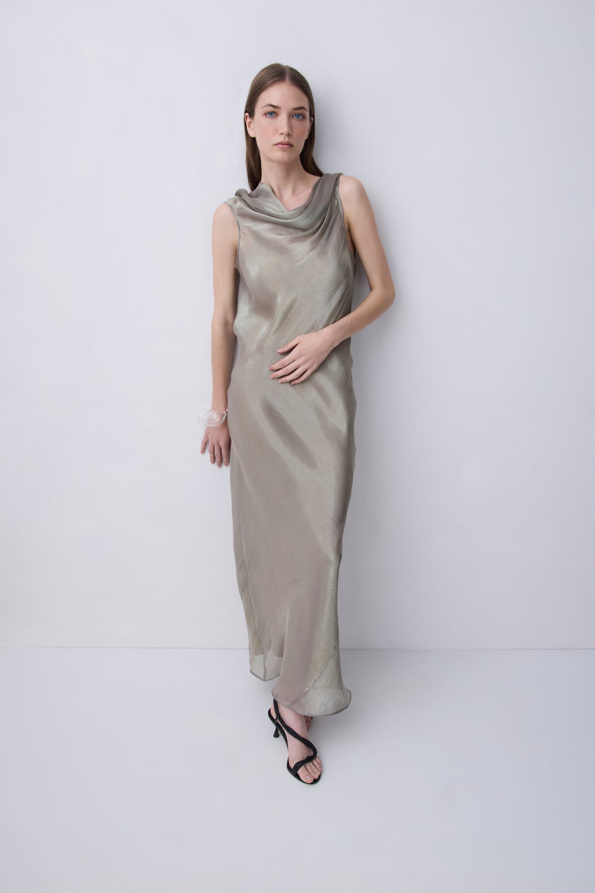 A woman modeling a sleeveless, cowl-neck maxi dress in a shimmering champagne-gold satin, standing against a plain light gray studio background.