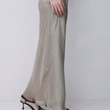 A model wearing a flowing, semi-sheer champagne beige maxi dress with a side slit, paired with black strappy high-heel sandals, shown from the waist down against a plain light gray studio background.