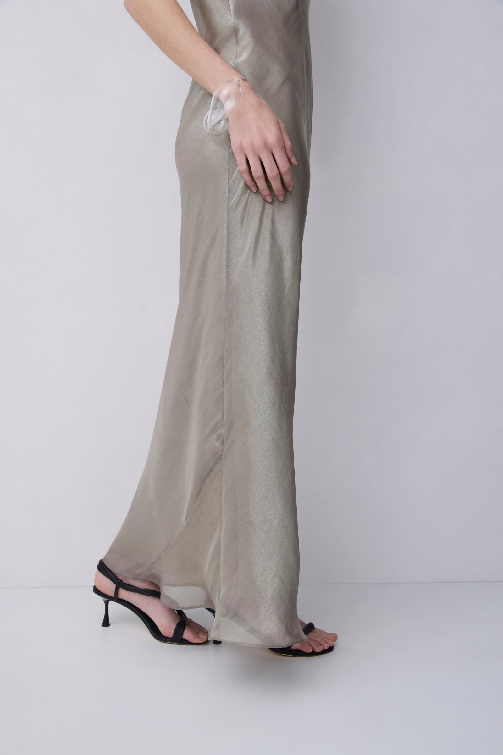 A model wearing a flowing, semi-sheer champagne beige maxi dress with a side slit, paired with black strappy high-heel sandals, shown from the waist down against a plain light gray studio background.
