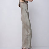A woman modeling a floor-length, sleeveless satin slip dress in a light taupe-beige color, paired with black strappy high-heeled sandals, set against a plain light gray studio background.