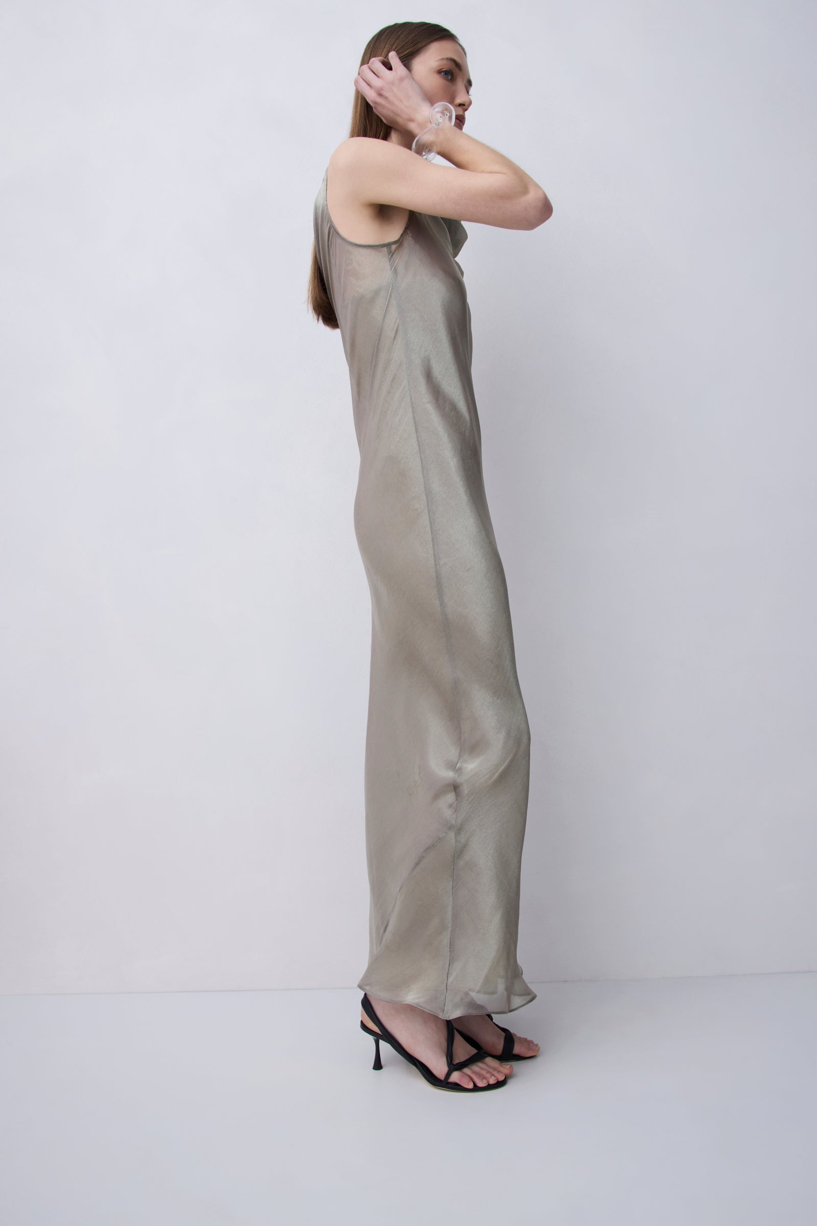 A woman modeling a floor-length, sleeveless satin slip dress in a light taupe-beige color, paired with black strappy high-heeled sandals, set against a plain light gray studio background.