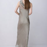 A woman modeling a sleeveless, floor-length draped dress in a light taupe-beige color, shown from the back against a plain light gray studio background.