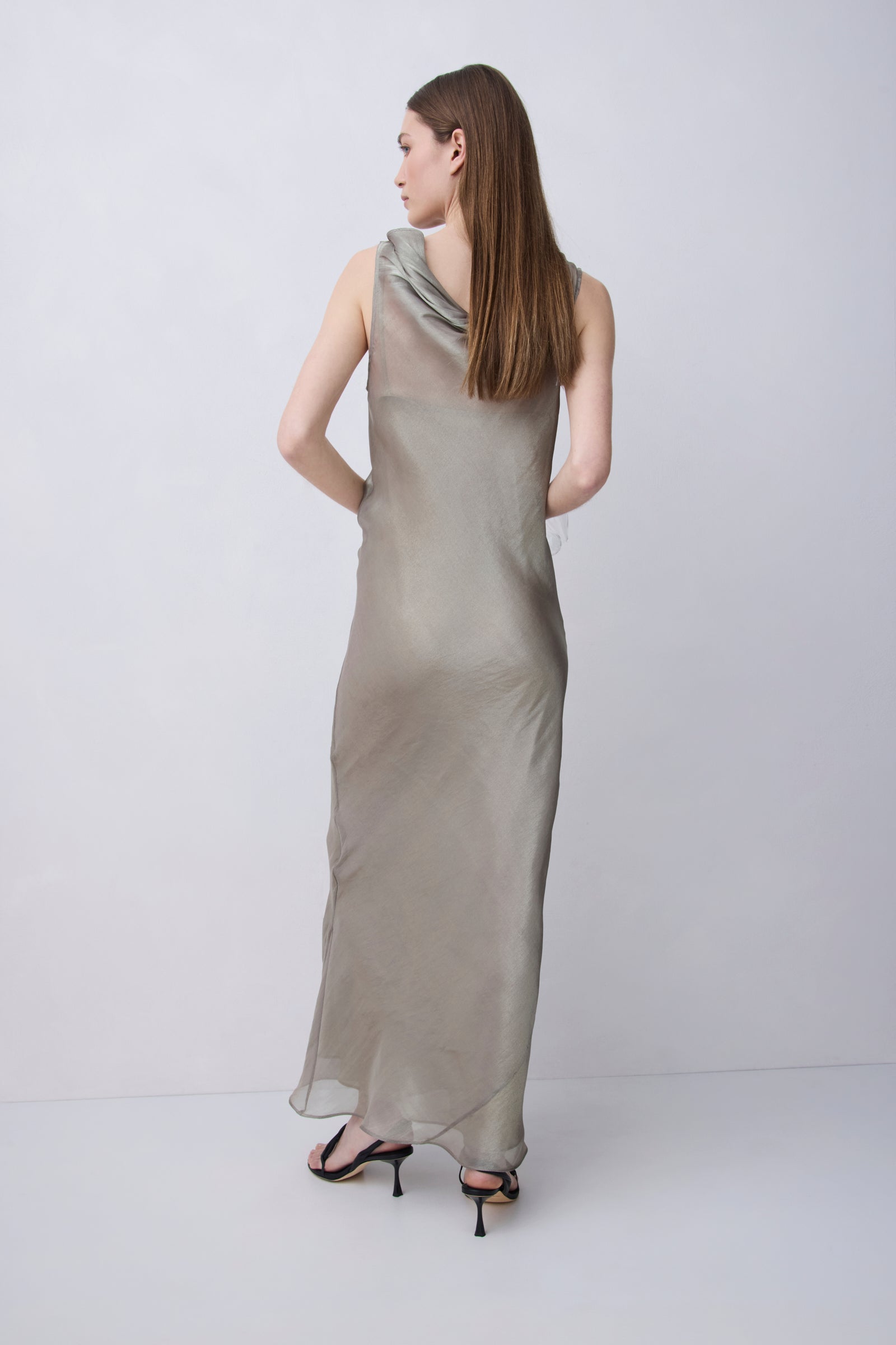 A woman modeling a sleeveless, floor-length draped dress in a light taupe-beige color, shown from the back against a plain light gray studio background.
