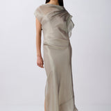 A model wearing a draped, semi-sheer floor-length dress in a light taupe-beige color, paired with gold strappy high-heeled sandals, set against a plain light gray studio background.