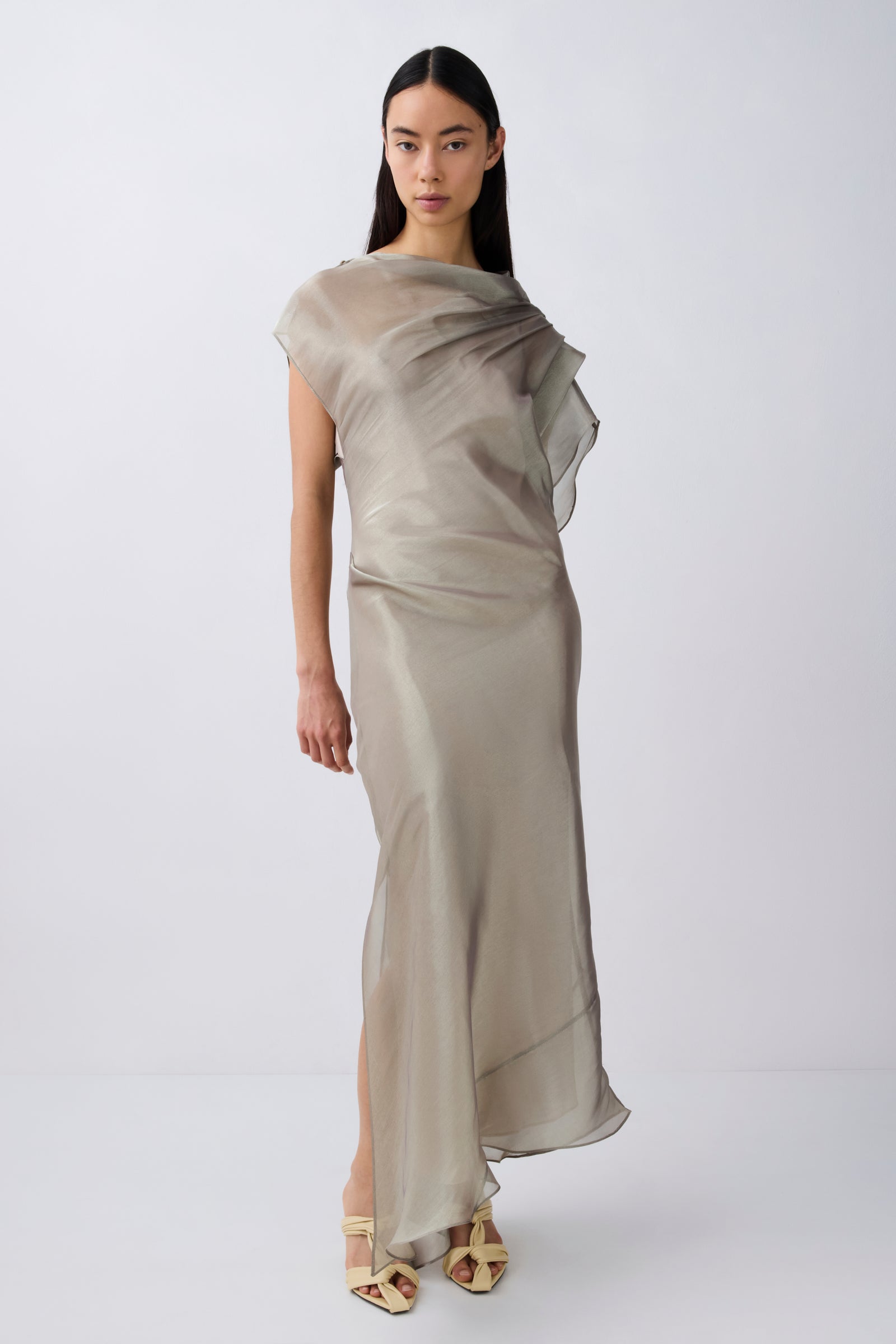 A model wearing a draped, semi-sheer floor-length dress in a light taupe-beige color, paired with gold strappy high-heeled sandals, set against a plain light gray studio background.
