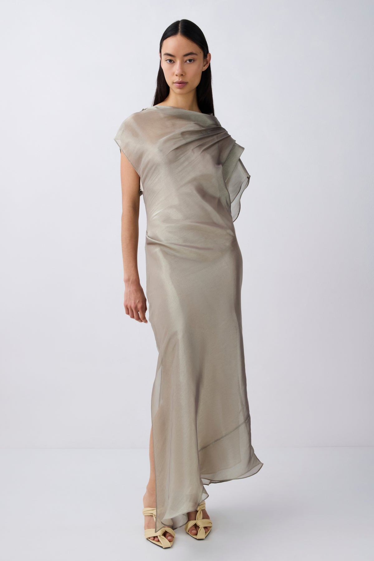 A model wearing a draped, semi-sheer floor-length dress in a light taupe-beige color, paired with gold strappy high-heeled sandals, set against a plain light gray studio background.