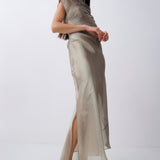 A model wearing a draped, one-shoulder satin maxi dress in a taupe-beige color, paired with light beige strappy heels, set against a plain light gray studio background.