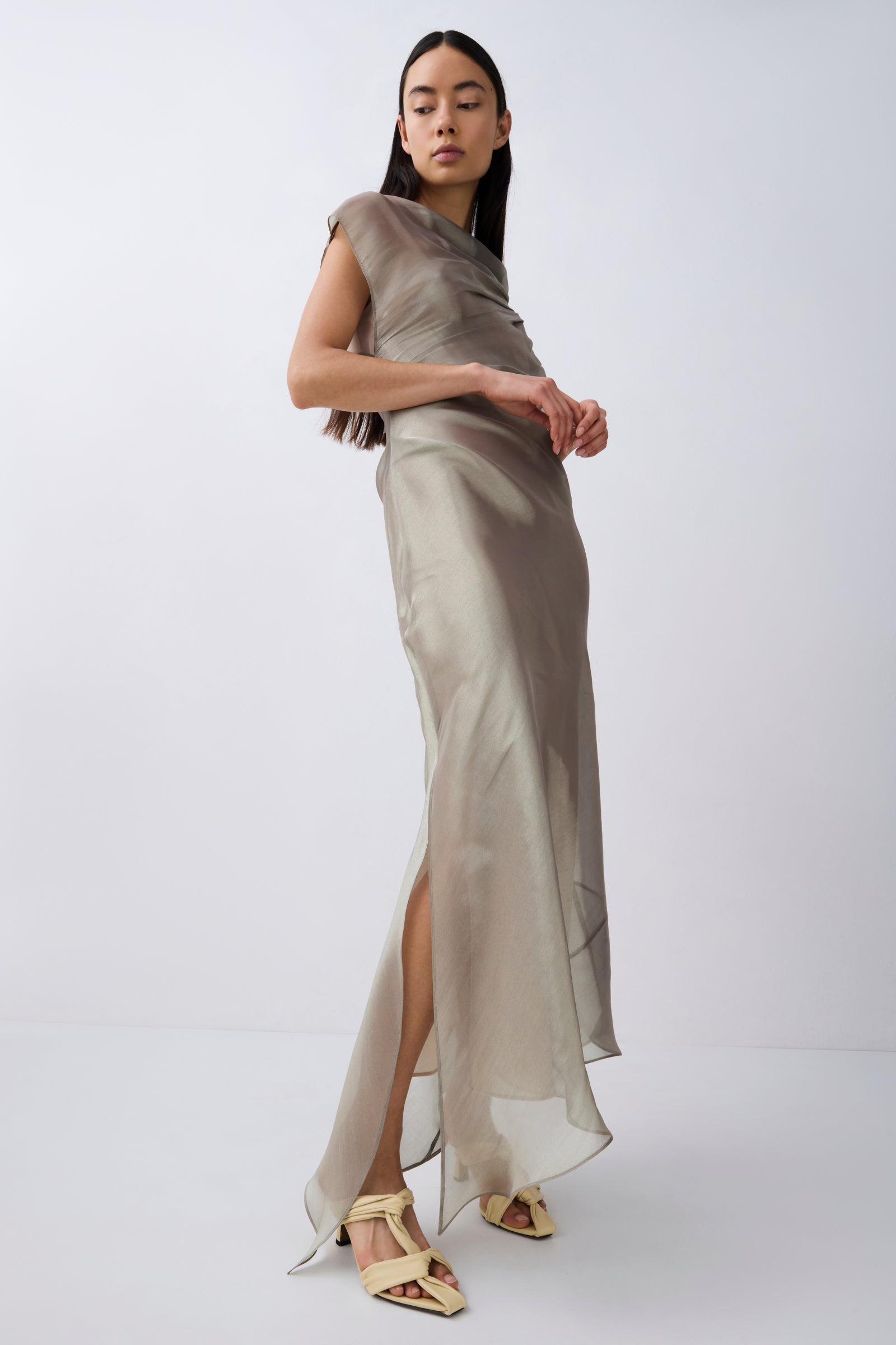 A model wearing a draped, one-shoulder satin maxi dress in a taupe-beige color, paired with light beige strappy heels, set against a plain light gray studio background.