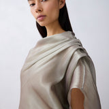 A woman with dark hair pulled back wears a draped, short-sleeved top in a light taupe-beige color with a subtle sheen, photographed against a neutral light gray background.