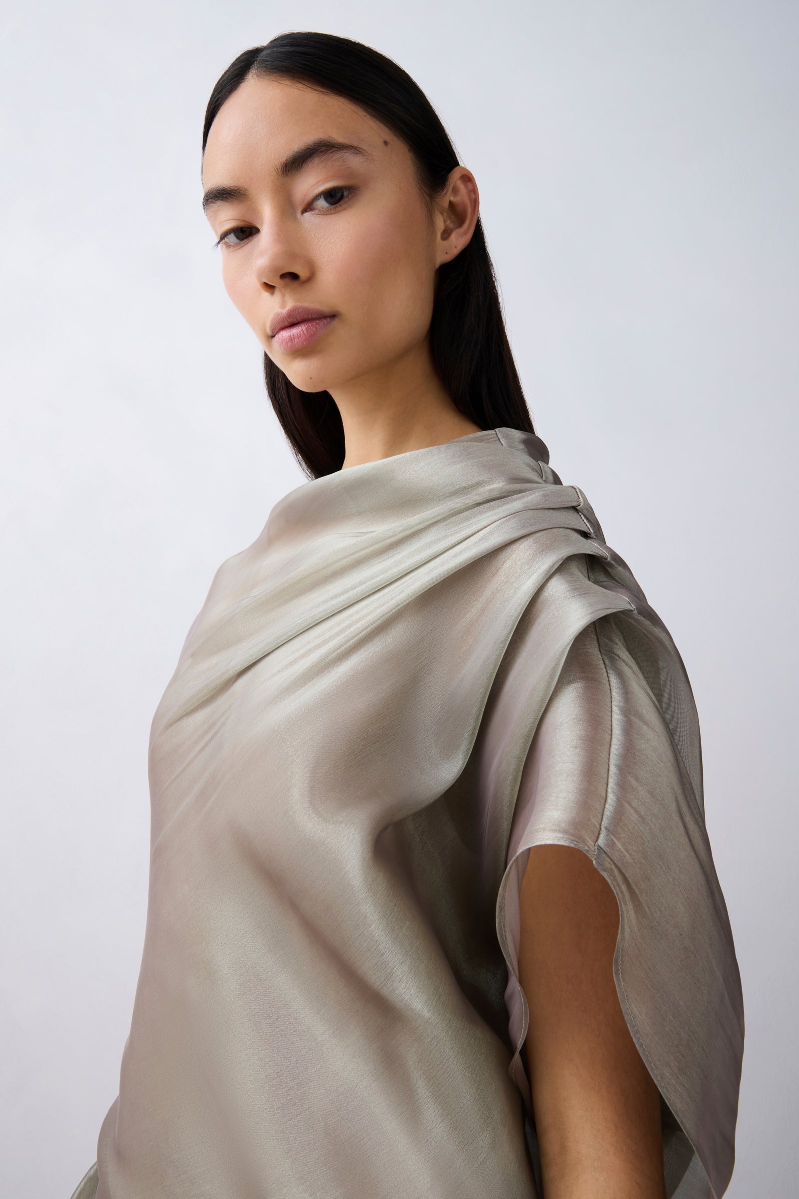 A woman with dark hair pulled back wears a draped, short-sleeved top in a light taupe-beige color with a subtle sheen, photographed against a neutral light gray background.