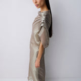 A model with long dark hair is shown from the side wearing a shimmering, metallic taupe-beige draped dress with short voluminous sleeves and a high gathered neckline, paired with light beige flat sandals, against a plain light gray studio background.