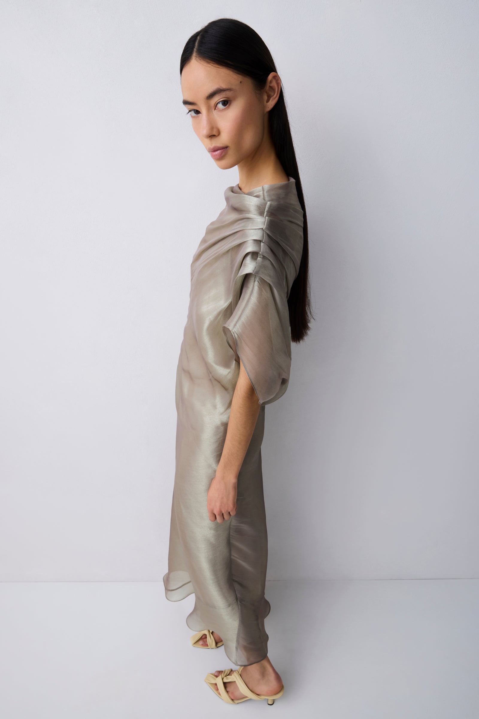 A model with long dark hair is shown from the side wearing a shimmering, metallic taupe-beige draped dress with short voluminous sleeves and a high gathered neckline, paired with light beige flat sandals, against a plain light gray studio background.