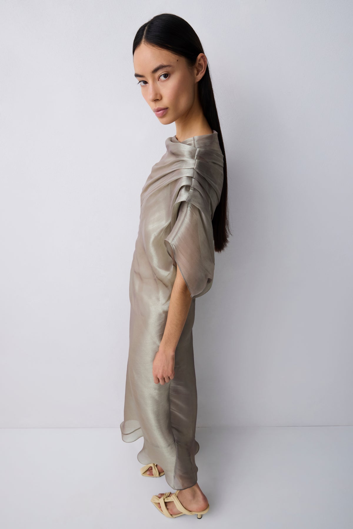 A model with long dark hair is shown from the side wearing a shimmering, metallic taupe-beige draped dress with short voluminous sleeves and a high gathered neckline, paired with light beige flat sandals, against a plain light gray studio background.