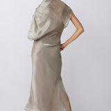 A woman shown from the back wearing a flowing, semi-sheer champagne beige maxi dress with a draped shawl collar, paired with beige high-heeled sandals, against a light gray studio background.