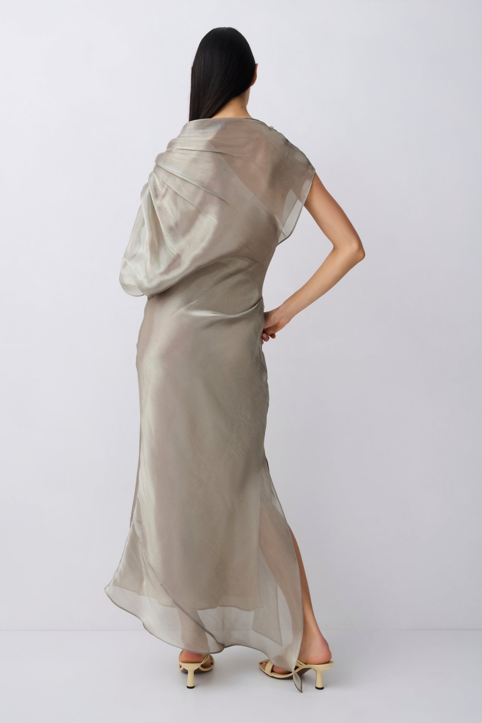 A woman shown from the back wearing a flowing, semi-sheer champagne beige maxi dress with a draped shawl collar, paired with beige high-heeled sandals, against a light gray studio background.
