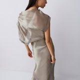 A woman modeling a floor-length draped dress in a light taupe-beige satin or silk-like fabric, photographed against a clean white studio background.