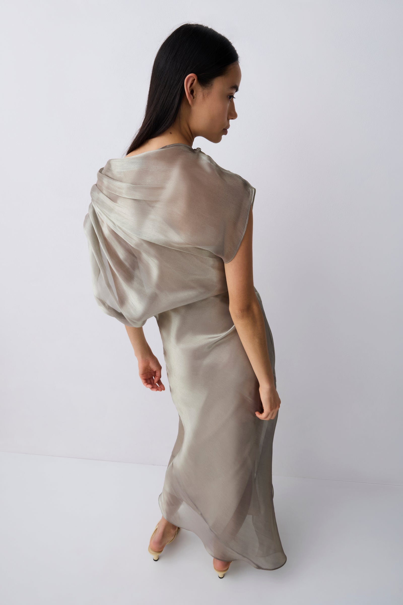 A woman modeling a floor-length draped dress in a light taupe-beige satin or silk-like fabric, photographed against a clean white studio background.