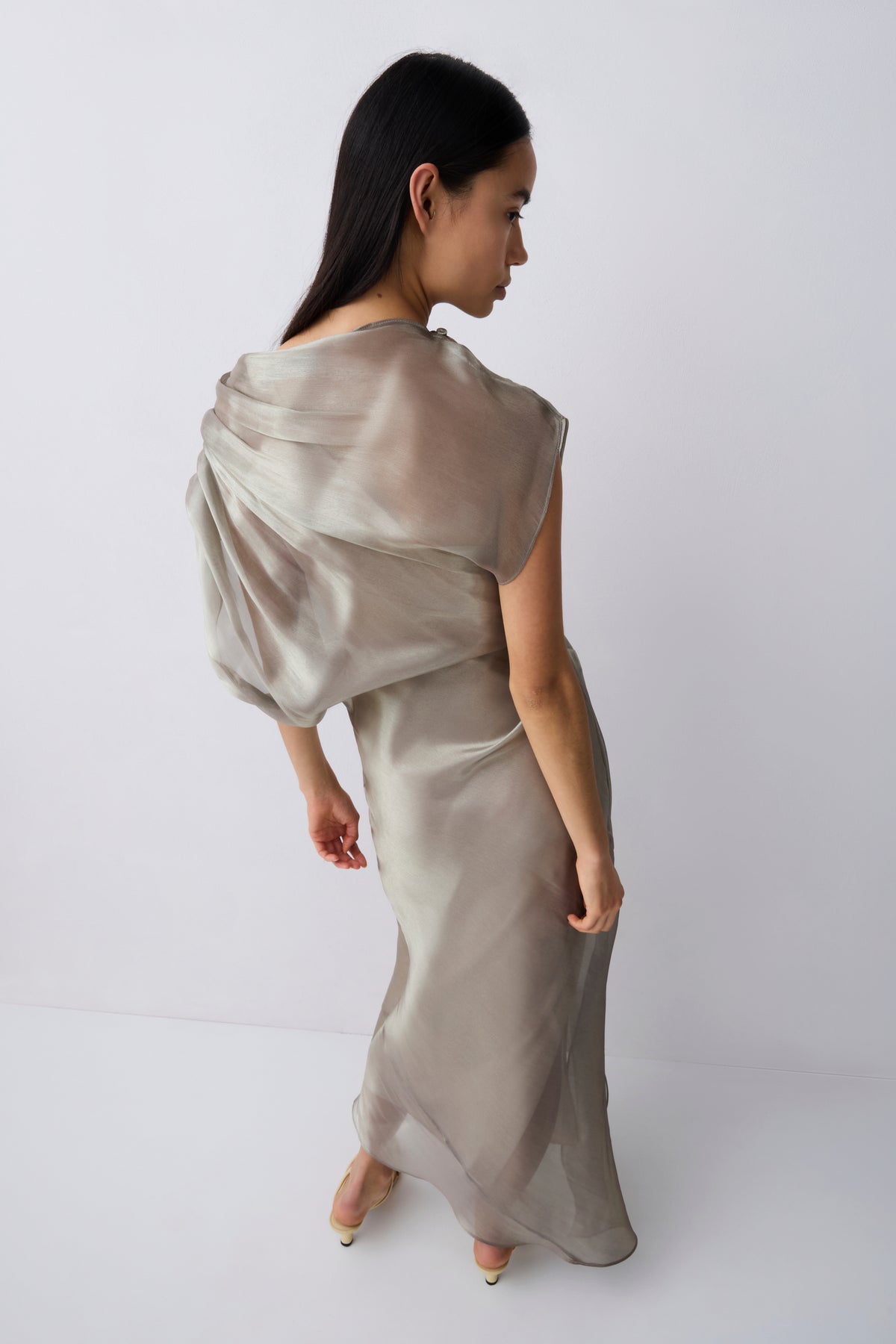 A woman modeling a floor-length draped dress in a light taupe-beige satin or silk-like fabric, photographed against a clean white studio background.